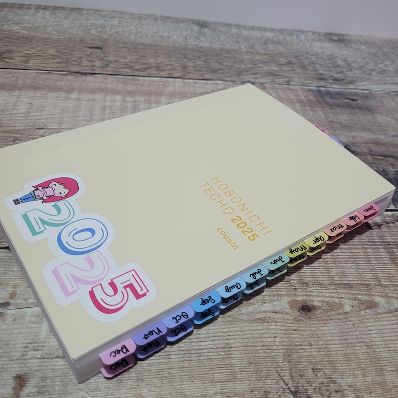 May include: A cream-colored Hobonichi Techo 2025 Cousin planner with colorful month tabs. The cover features a sticker with the year 2025 and a cartoon character. The planner is open, revealing the month tabs.
