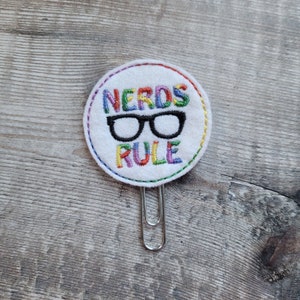 May include: A white round fabric bookmark with rainbow colored embroidery that reads "NERDS RULE" with a pair of black glasses in the center. The bookmark is attached to a silver metal paper clip.