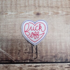 Può includere: Una graffetta a forma di cuore in feltro bianco con le parole "Fuck Off" ricamate con filo rosso. Il cuore è contornato da filo viola e attaccato a una graffetta d'argento. Lo sfondo è una superficie di legno.