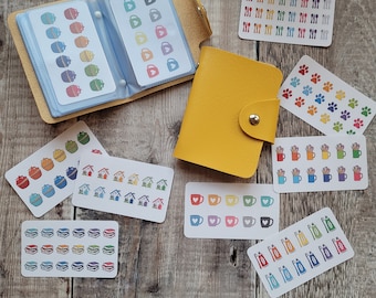 May include: Assortment of colorful stickers featuring various designs, including cupcakes, houses, coffee cups, books, and paw prints. The stickers are arranged on a wooden surface with a yellow leather notebook and a clear plastic sticker book.