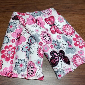 May include: A pair of pink and gray fabric heat packs with a butterfly and floral pattern. The heat packs are sewn together and have a white background.