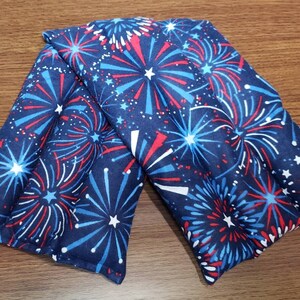 May include: Two blue fabric heat packs with a red, white, and blue firework pattern. The heat packs are folded in half and are lying on a wooden surface.
