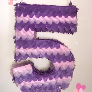 May include: A large, purple and pink number five piñata, decorated with layered paper in a scalloped pattern. The piñata is hanging from a white string. The text "By Piñataland" is visible at the bottom.