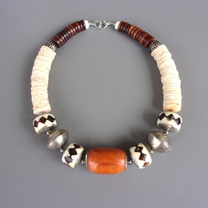 Op de afbeelding: Een ketting met een centrale gladde oranje tonvormige kraal, geflankeerd door zilverkleurige metalen bollen en witte kralen met donkerbruine zigzagpatronen. De bovenste delen bestaan uit gestapelde witte schijfkralen en donkerbruine houten schijfkralen.