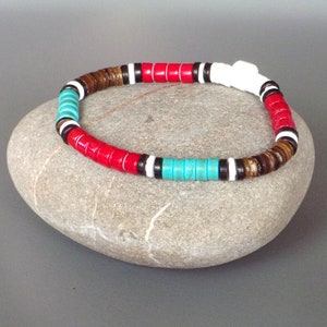 Op de afbeelding: Een veelkleurige kralen armband met rode, witte, bruine, zwarte en turquoise kralen. De armband ligt op een grijze steen.