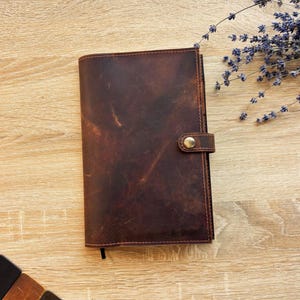 May include: A brown leather journal with a brass clasp, closed and lying flat on a light wood surface. The leather has a distressed appearance. A sprig of lavender is in the background.