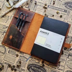 Peut inclure: Un journal en cuir marron ouvert avec un carnet noir et trois stylos. Le journal a un intérieur beige clair. Le carnet porte le texte "Axent" et "Partner Notebook". Une plume et un encrier sont visibles en arrière-plan.
