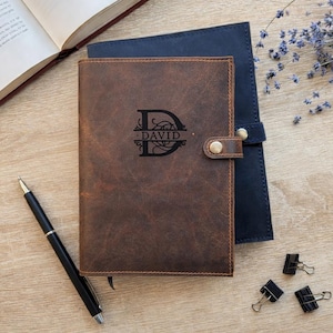 May include: A brown leather journal with the name "DAVID" engraved on the cover, secured with a snap closure. A black pen and binder clips are next to the journal. A blue journal is behind the brown one.