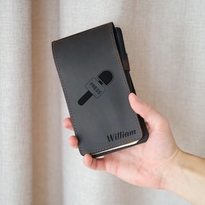 Puede incluir: Una funda de cuero gris oscuro para cuaderno con un gráfico de micrófono y la palabra "PRESS" impresa. El nombre "William" está grabado en la parte inferior. La funda se sostiene en una mano.