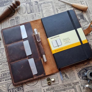 May include: A leather notebook cover with a black Moleskine notebook and a pen inside. The cover has card slots and is brown. A leather crafting tool and other tools are also visible.