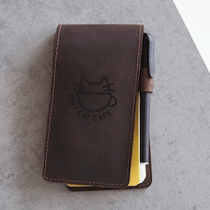 May include: A dark brown leather notepad holder with a pen and yellow notepad. The holder features the text "Tiny Cat Cafe" with a cat and coffee cup logo. The notepad holder is closed and has a stitched border.