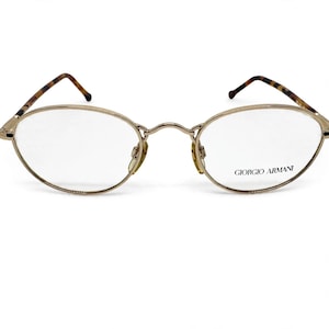 May include: Oval-shaped eyeglasses with a thin gold metal frame and clear lenses. The right lens features the text GIORGIO ARMANI printed in black. The temples are thin with a brown and black tortoiseshell pattern at the tips.