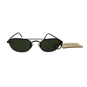 May include: Black oval-shaped sunglasses with dark green lenses. The sunglasses have a thin, black metal frame and a small tag attached with the text "Calvin Klein" printed on it. The sunglasses are a classic style.
