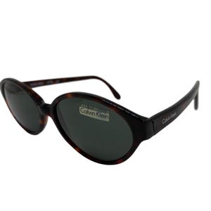 May include: Oval-shaped sunglasses with a tortoiseshell pattern frame and dark lenses. The Calvin Klein logo is visible on the lens and arm. The sunglasses are a classic accessory.