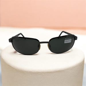 May include: Black oval sunglasses with a thin black frame. The sunglasses have dark lenses and a small white sticker on the right lens with the text "100% UV PROTECTION OLIVER". The sunglasses are displayed on a white surface.