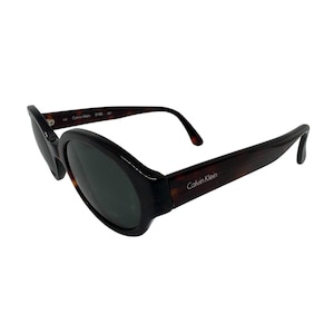 May include: Dark brown Calvin Klein sunglasses with oval lenses. The frames have a tortoiseshell pattern and the Calvin Klein logo is visible on the temples. The sunglasses are angled slightly, showing the shape and design.