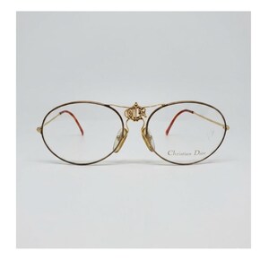May include: Gold-tone metal eyeglasses with oval lenses and a bridge detail. The frames have a thin, delicate design with red accents on the temples. The brand name "Christian Dior" is printed on the right lens.