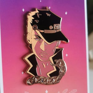 May include: A metal enamel pin featuring a stylised character wearing a black cap and jacket with a gold chain, mounted on a rectangular card with a purple to orange gradient background, the Roman numeral XVII, and the text THE STAR.