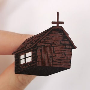 Puede incluir: Una pequeña cabaña de madera marrón con una cruz en el techo. La cabaña tiene una sola ventana y una puerta. La cabaña está recortada en papel y está siendo sostenida por un dedo.