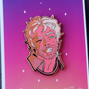 Op de afbeelding: Een roze en witte emaille pin met een cartoonillustratie van een man met een baard en een groen oog. De pin is geplaatst tegen een roze en paarse gradiëntachtergrond met de tekst "IX THE HERMIT" onderaan.