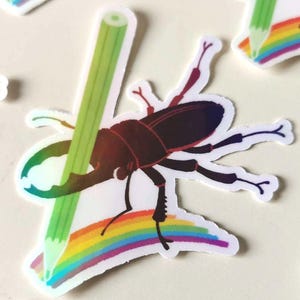 Puede incluir: Una colección de pegatinas troqueladas que muestran un ciervo volante de color marrón oscuro con grandes mandíbulas sobre un arcoíris vibrante. Cada escarabajo sostiene un lápiz verde claro que se extiende diagonalmente por el diseño.
