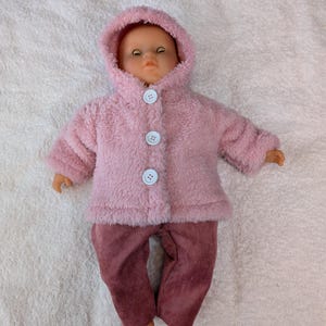 May include: A doll dressed in a pink hooded jacket with three white buttons and corduroy trousers. The jacket is made of a soft, fluffy material, and the trousers are a muted rose colour. The doll is on a white, textured surface.