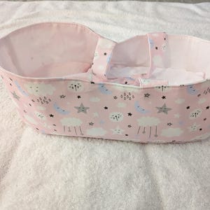 May include: A pink doll bassinet with a white interior and handles. The exterior features a pattern of clouds, moons, and stars in blue, gray, and black. The bassinet is designed for a doll.