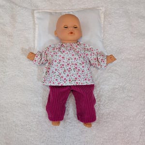 Peut inclure: Une poupée pour bébé vêtue d'un haut à imprimé floral et d'un pantalon en velours côtelé magenta, reposant sur un oreiller blanc. La poupée a la tête chauve et est sur une surface blanche texturée.