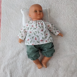 Peut inclure: Une poupée vêtue d'un haut blanc à imprimé floral et d'un short vert, reposant sur un petit oreiller blanc. La poupée est posée sur une serviette blanche. Le haut a des manches longues et une encolure ronde. Le short est de couleur verte unie.