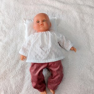 Könnte beinhalten: Eine Vintage-Babypuppe mit einem weißen Lochmuster-Oberteil und einer bordeauxfarbenen Cordhose. Die Puppe liegt auf einem kleinen weißen Kissen auf einer weißen, strukturierten Oberfläche. Die Puppe hat einen kahlen Kopf und geschlossene Augen.