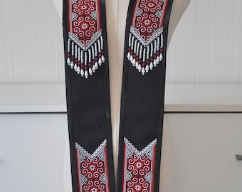 May include: A black stole featuring intricate red, white, and black geometric embroidery patterns. The garment includes beaded fringe accents in white and red at the ends of the panels. The text CROSSROADS BY NANCY is printed at the bottom.
