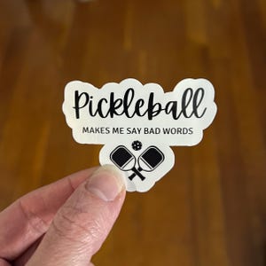 May include: White sticker with black text that reads "Pickleball Makes Me Say Bad Words". The sticker also features a black and white illustration of two pickleball paddles and a ball.