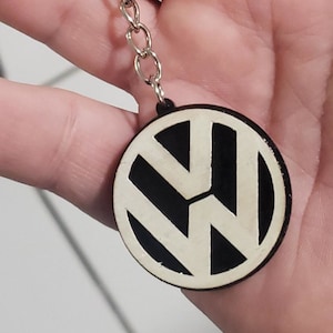 May include: A round keychain featuring the Volkswagen logo. The logo is a white design on a black background. The keychain is attached to a silver chain.