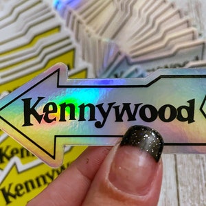 K&ouml;nnte beinhalten: Ein holografischer Aufkleber mit einem nach links zeigenden Pfeil und dem Text "Kennywood" in schwarzen Buchstaben.