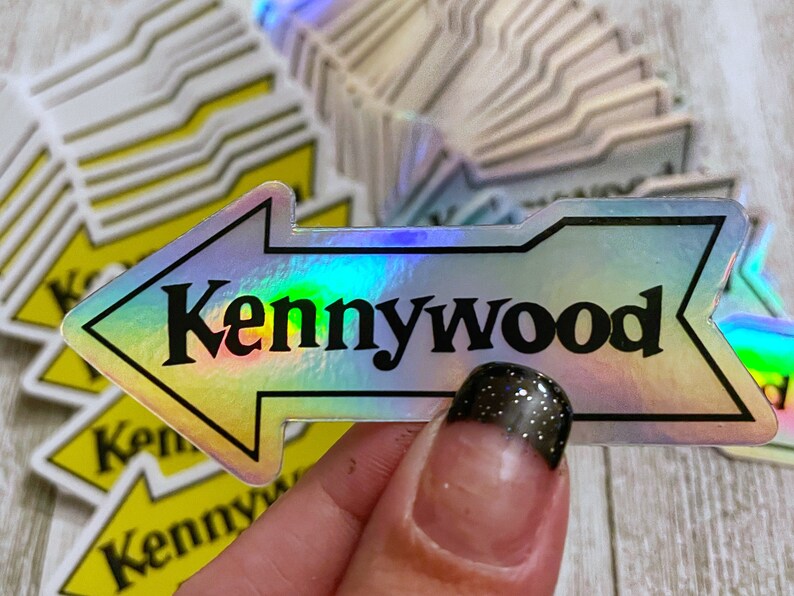 K&ouml;nnte beinhalten: Ein holografischer Aufkleber mit einem nach links zeigenden Pfeil und dem Text "Kennywood" in schwarzen Buchstaben.