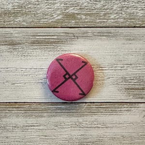 May include: A round, magenta button with a black geometric design. The design features two intersecting lines forming an X shape, with a right angle symbol in the center. The button is set against a light wood grain background.