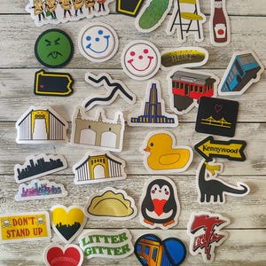 May include: A collection of colorful stickers with various designs, including smiley faces, a pickle, a penguin, and cityscapes. The stickers feature text like "Yinz," "Heinz," and "Kennywood." The stickers are arranged on a light wood surface.