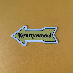 K&ouml;nnte beinhalten: Ein gelber Pfeil-Aufkleber mit wei&szlig;em Rand und dem Text "Kennywood" in Schwarz.