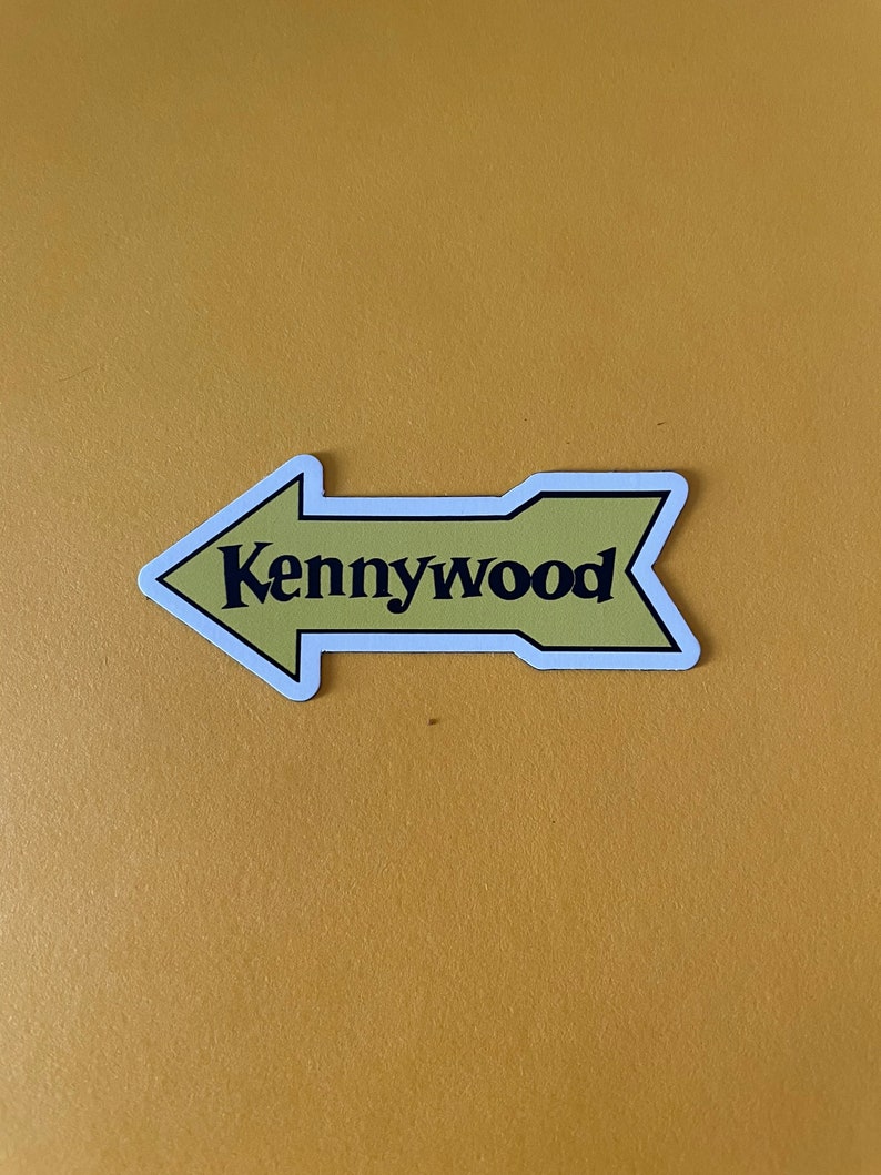 K&ouml;nnte beinhalten: Ein gelber Pfeil-Aufkleber mit wei&szlig;em Rand und dem Text "Kennywood" in Schwarz.
