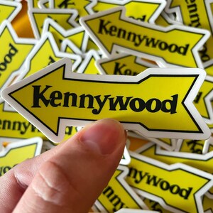 K&ouml;nnte beinhalten: Ein gelber Pfeil-Aufkleber mit dem Text "Kennywood" in schwarzen Buchstaben. Der Aufkleber wird von einer Hand gehalten und im Hintergrund befinden sich viele weitere &auml;hnliche Aufkleber.
