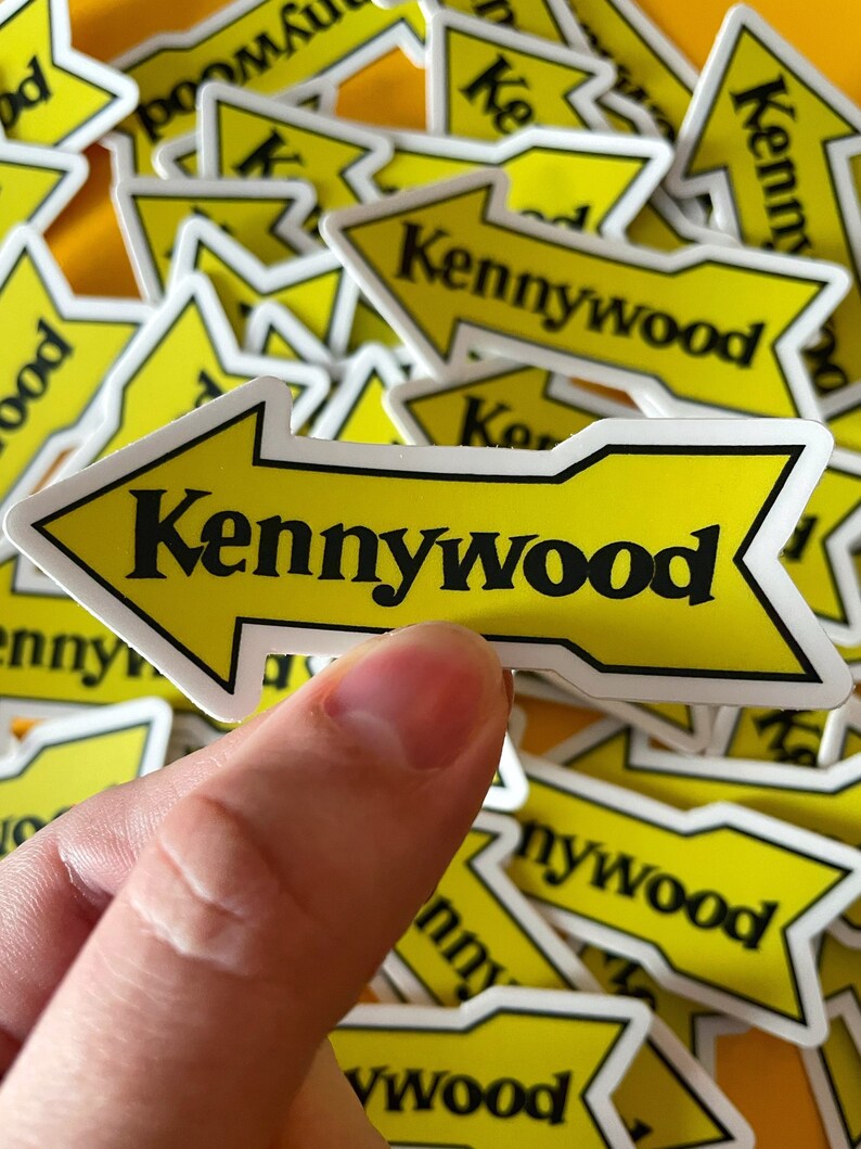 K&ouml;nnte beinhalten: Ein gelber Pfeil-Aufkleber mit dem Text "Kennywood" in schwarzen Buchstaben. Der Aufkleber wird von einer Hand gehalten und im Hintergrund befinden sich viele weitere &auml;hnliche Aufkleber.