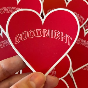 K&ouml;nnte beinhalten: Ein Stapel roter herzf&ouml;rmiger Aufkleber mit wei&szlig;em Rand. Der oberste Aufkleber tr&auml;gt den Schriftzug "GOODNIGHT" in wei&szlig;en umrandeten Buchstaben.