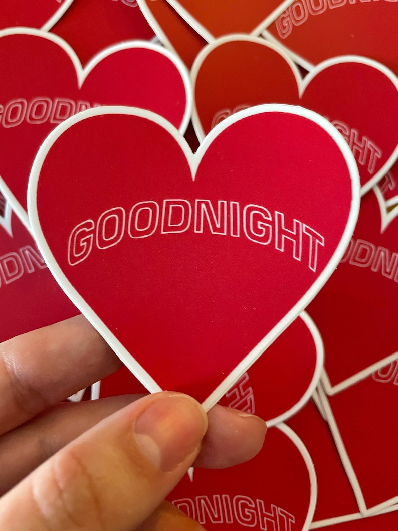 K&ouml;nnte beinhalten: Ein Stapel roter herzf&ouml;rmiger Aufkleber mit wei&szlig;em Rand. Der oberste Aufkleber tr&auml;gt den Schriftzug "GOODNIGHT" in wei&szlig;en umrandeten Buchstaben.