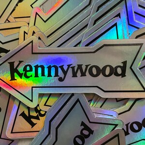 K&ouml;nnte beinhalten: Ein Stapel von regenbogenfarbenen silbernen Pfeil-Aufklebern mit dem Text "Kennywood" in schwarzen Buchstaben.