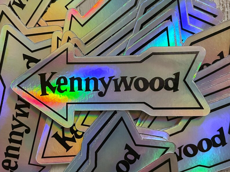 K&ouml;nnte beinhalten: Ein Stapel von regenbogenfarbenen silbernen Pfeil-Aufklebern mit dem Text "Kennywood" in schwarzen Buchstaben.