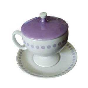 May include: A white ceramic teacup with a purple lid and a white saucer. Both the cup and saucer have a purple polka dot pattern.