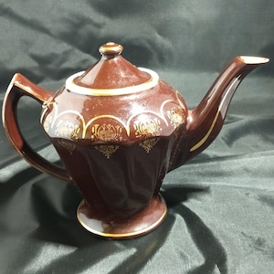 May include: A brown teapot with gold accents and a white rim. The teapot has a rounded body and a spout on the side. The teapot is sitting on a black surface.