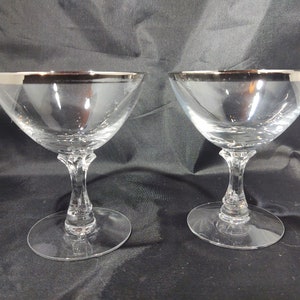 Peut inclure: Deux coupes à champagne transparentes avec des bords argentés. Les verres ont un design classique avec un bol large et peu profond et une longue tige. La base est ronde et plate. Les verres sont probablement en verre.