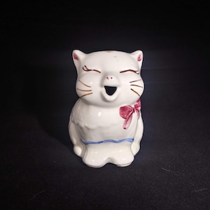 Peut inclure: Pichet en céramique blanche en forme de chat avec un nœud rouge et une bordure bleue. Le chat a des moustaches peintes en brun et les yeux fermés avec la bouche ouverte.