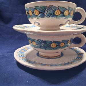 Puede incluir: Dos tazas de té y platillos de cerámica apilados. Las tazas y los platillos son de color blanquecino con un diseño pintado a mano de hojas azules y frutas amarillas. Los bordes y las asas son redondeados.